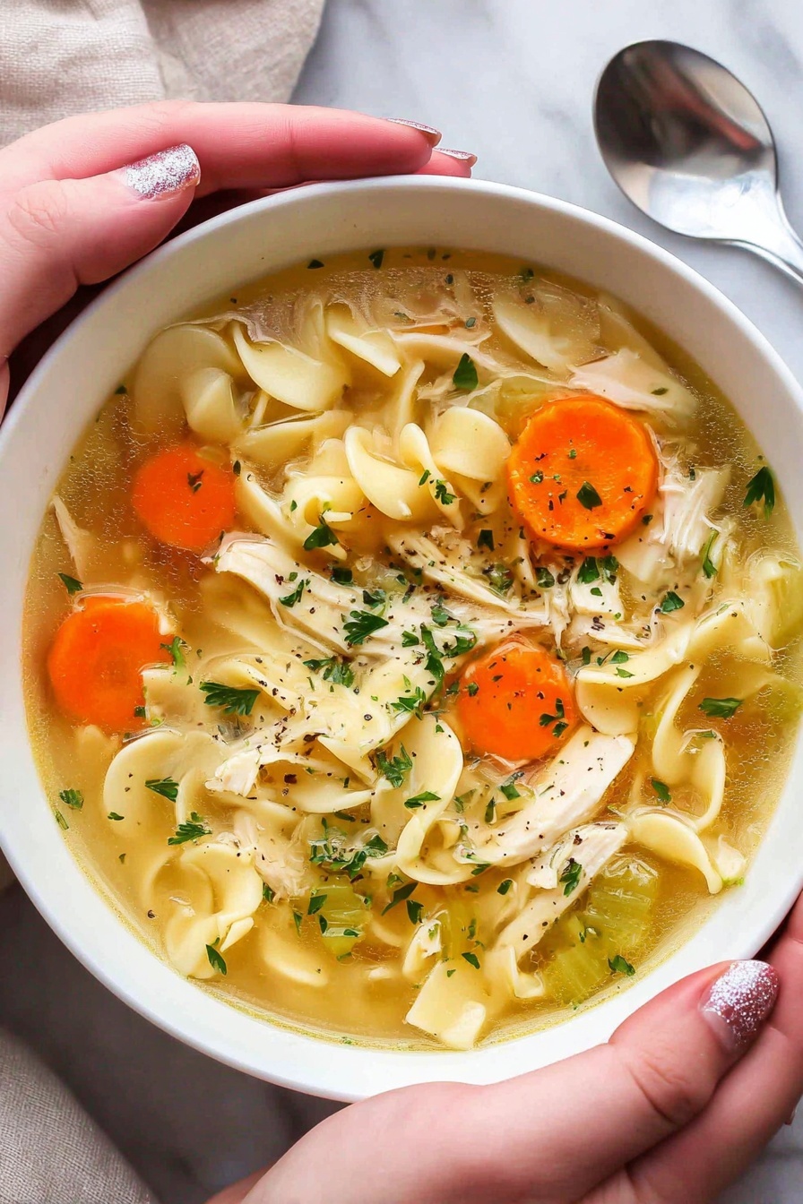 A white bowl filled with chicken noodle soup is held by two woman's hands with glittery pink nail polish. The soup has a clear yellow broth with pieces of shredded chicken, wide egg noodles, round slices of orange carrots, and small green celery chunks. Fresh green parsley flakes float on the surface, adding a touch of color. The bowl is placed on a white marbled surface with a silver spoon partly visible on the side. The image has a warm and cozy feel. photo taken with an iphone --ar 2:3 --v 7