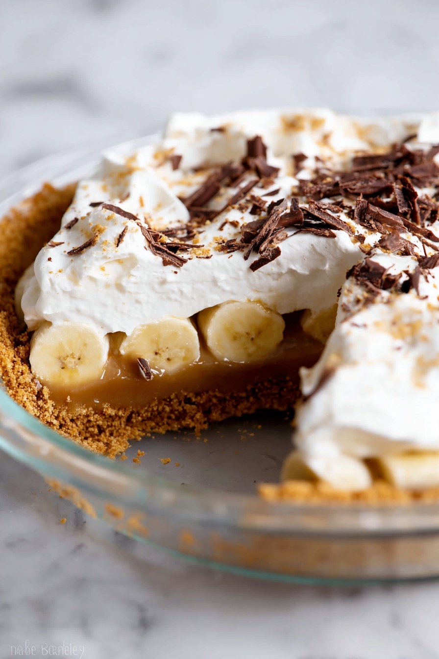 The image shows a three-layered dessert in a clear glass pie dish on a white marbled surface. The bottom layer is a crumbly, golden-brown crust. Above this is a thick layer of banana slices covered by a caramel-like filling. The top layer consists of a thick, fluffy white whipped cream with dark brown chocolate shavings sprinkled over it. A slice has been taken out, revealing the layers clearly. Photo taken with an iphone --ar 2:3 --v 7