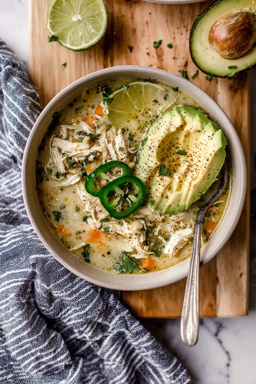 A bowl of creamy chicken soup with visible shredded chicken and small pieces of orange vegetables floating in a light broth, topped with three slices of green jalapeno, half a lime placed on the soup’s edge, and three thin slices of avocado sprinkled with black pepper and fresh green herbs. The bowl is light beige and placed on a wooden board with two halves of squeezed lime nearby and scattered green herbs around. A silver spoon rests inside the bowl on the right side, and a navy blue and white striped cloth lies underneath. The background features a white marbled surface. Photo taken with an iphone --ar 2:3 --v 7