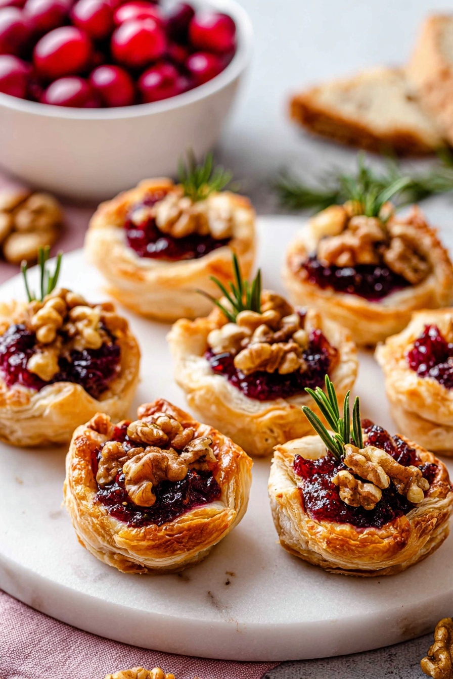 The image shows seven small, puffed pastry tarts arranged on a white marbled surface. Each tart has a golden-brown, flaky crust folded up around two layers inside: a deep red jam or fruit spread at the bottom and topped with crushed walnuts and whole walnut pieces. A small sprig of green rosemary sits on top of each tart, adding a fresh touch. In the background, there is a white square bowl filled with bright red cherries, and part of a sliced bread loaf is also visible on the left side. The setup is bright and inviting with a focus on the texture and color contrast of the tarts. photo taken with an iphone --ar 2:3 --v 7