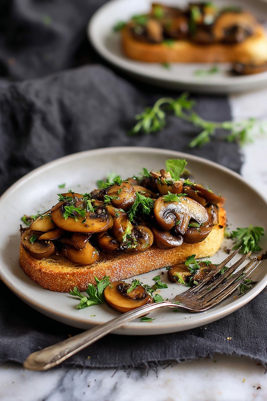 The image shows a white plate holding a single thick slice of lightly toasted bread with a golden-brown crust as the base layer. On top of the bread is a generous layer of sautéed mushroom slices, cooked to a dark golden color with glossy texture, arranged unevenly. Fresh green finely chopped herbs are sprinkled over the mushrooms and plate adding fresh color. A silver fork rests on the plate’s edge. The plate is placed on a soft, dark gray cloth over a white marbled surface. Another similar white plate with the same dish is blurred in the background. Photo taken with an iphone --ar 2:3 --v 7