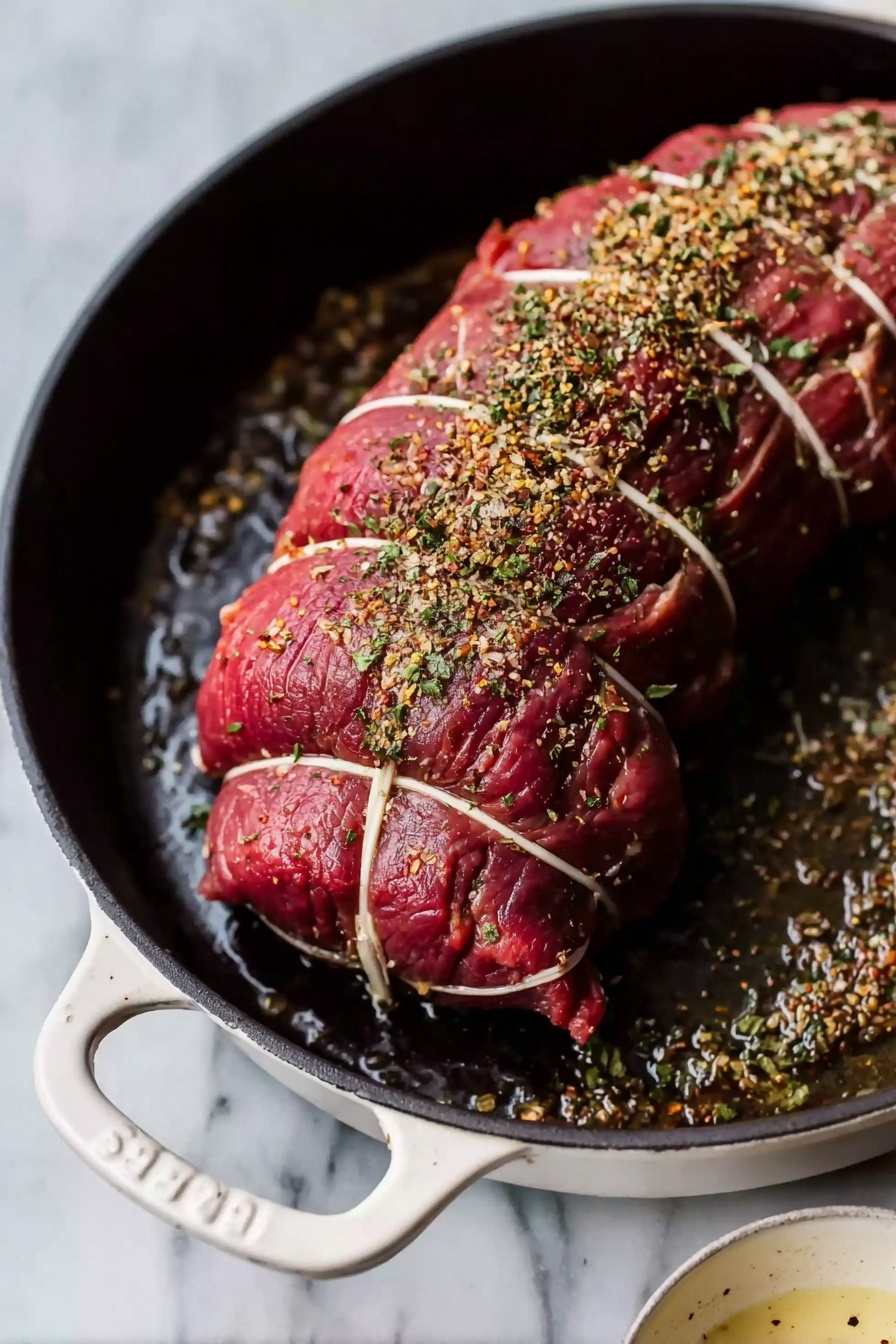 A thick piece of raw meat shaped in a curved cylinder is placed inside a white cast iron pan with a black inside surface. The meat is tied with thin white strings wrapped around it, and it is covered with small dried herbs and coarse salt sprinkled evenly on top. The pan sits on a white marbled surface with a small white bowl of creamy sauce nearby. Photo taken with an iphone --ar 2:3 --v 7