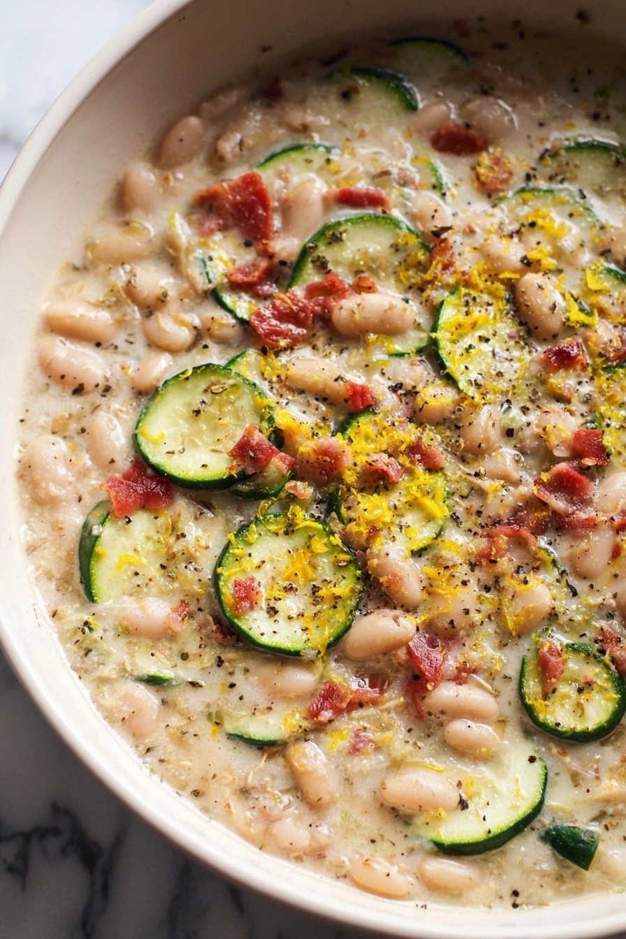 The image shows a close-up of a creamy dish in a white pot with a black rim, filled with small white beans, thin round slices of green zucchini, small pieces of crispy bacon in reddish-brown, and finely grated yellow lemon zest scattered on top. The soup-like base is light beige and smooth with visible flecks of black pepper and herbs sprinkled evenly throughout. The dish looks thick and hearty, with a mix of soft and slightly crunchy textures from the vegetables and bacon. The background is a white marbled surface with soft natural lighting. photo taken with an iphone --ar 2:3 --v 7