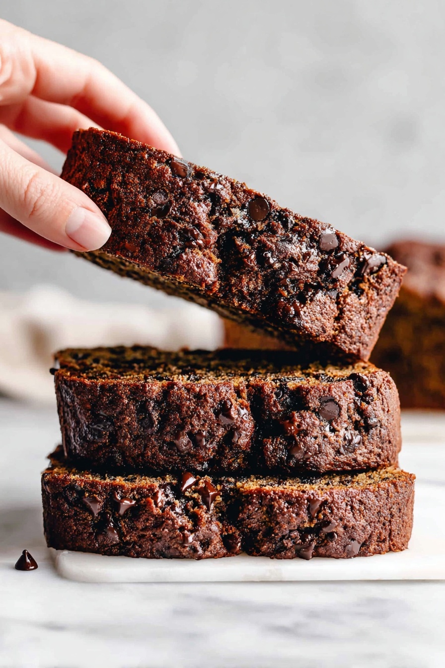 The image shows three thick slices of chocolate chip banana bread stacked on a white marbled surface. The bottom two slices lie flat with a dark brown color and many small chocolate chips scattered throughout, giving a chunky texture. A woman's hand is gently lifting the top slice, showing its moist interior with a mix of light brown banana bread and melted chocolate spots. The background includes blurred pieces of bread, keeping the focus on the front stack. Photo taken with an iphone --ar 2:3 --v 7