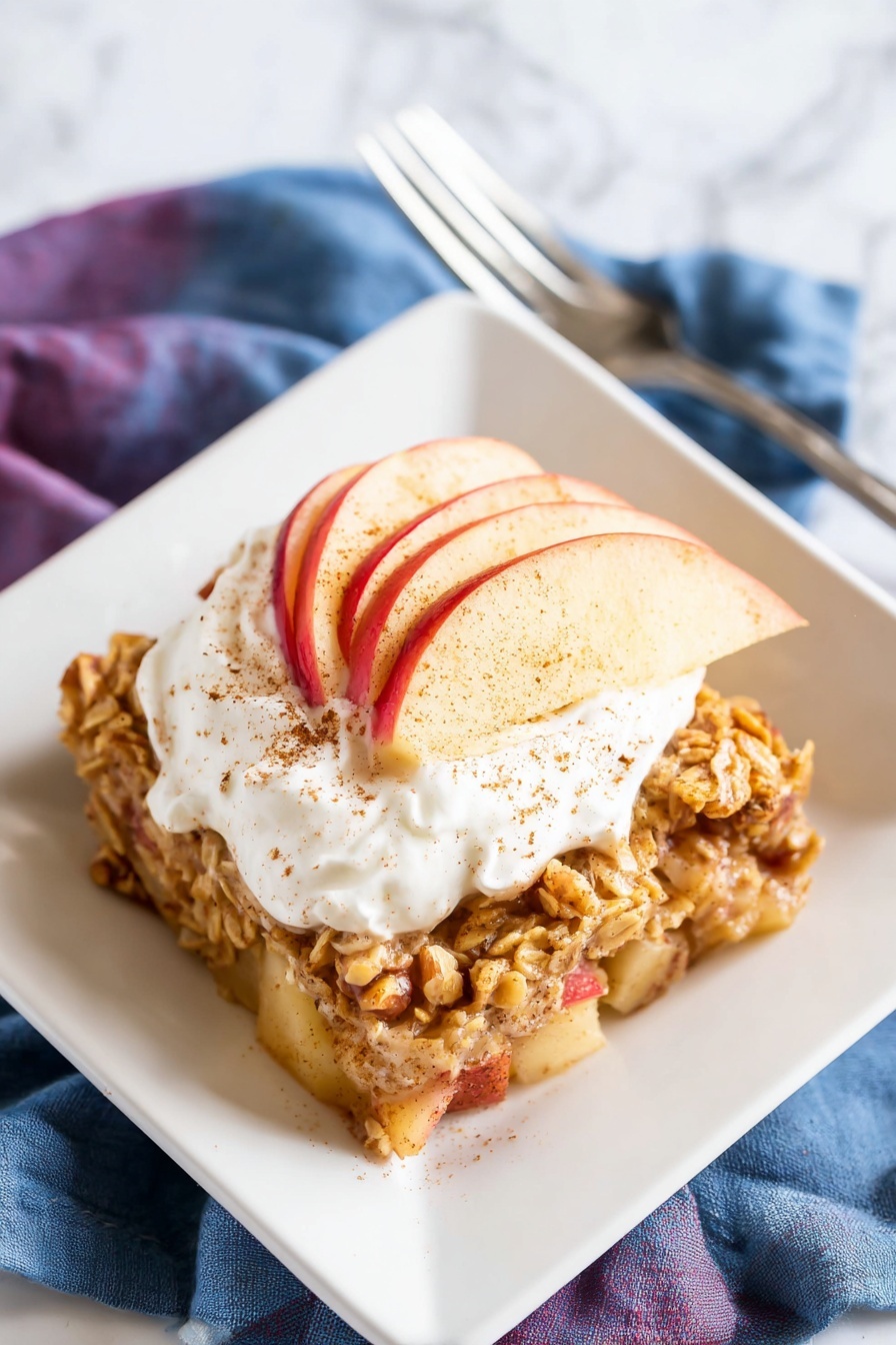 A square white plate holds a single thick slice of baked oat and apple mixture, showing soft chunks of apple with golden brown baked oats and nuts mixed throughout. On top, there is a dollop of white whipped cream, sprinkled lightly with brown cinnamon powder. Thin red-skinned apple slices fan out slightly under the cream, adding a fresh touch. The plate rests on a soft blue and purple cloth napkin, all placed on a white marbled surface. Photo taken with an iphone --ar 2:3 --v 7