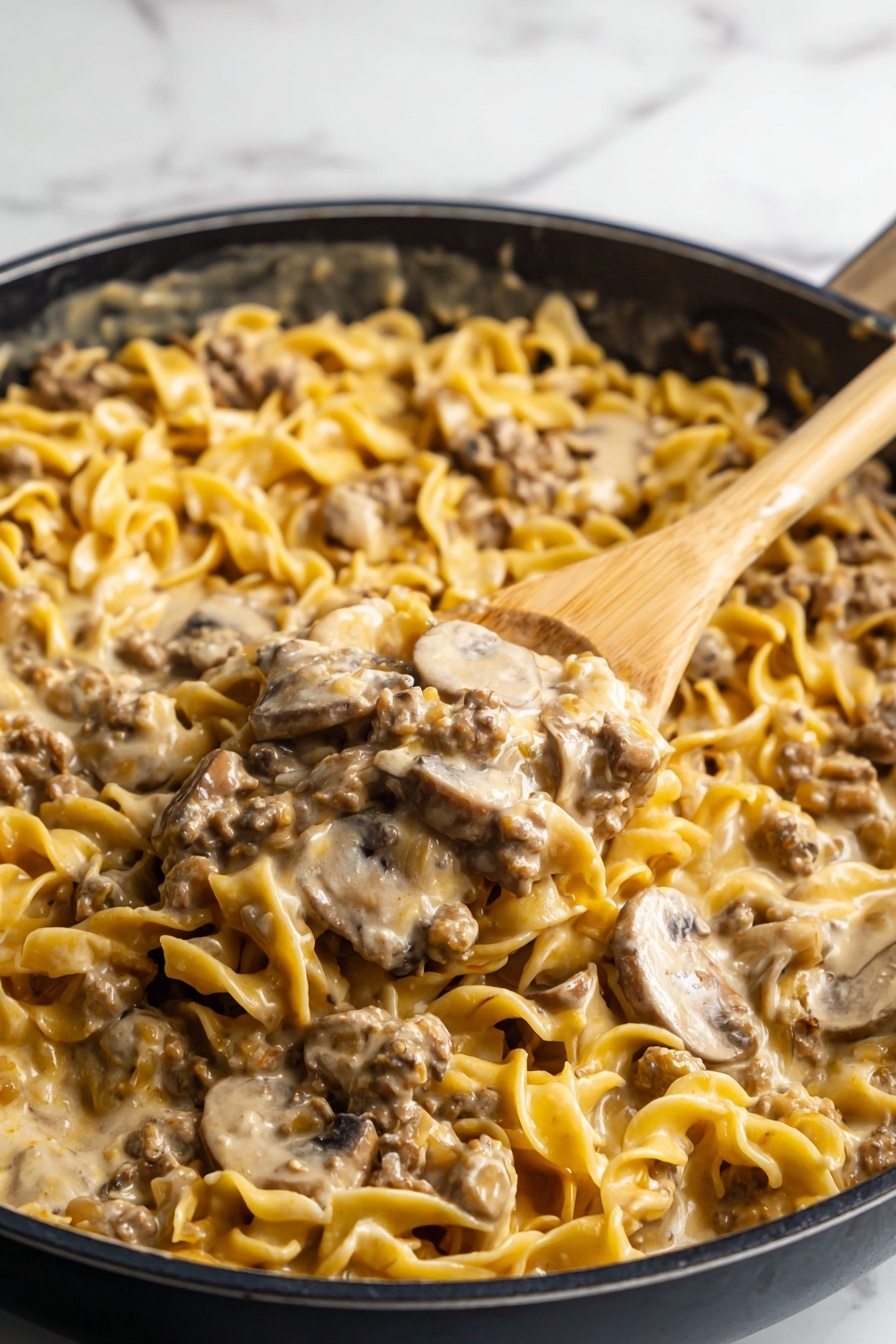 The image shows a close-up of a black skillet filled with creamy noodles mixed with small pieces of cooked ground meat and sliced mushrooms. The noodles are broad and curly with a pale yellow color, coated in a thick light beige sauce that gives the dish a moist texture. The ground meat is scattered evenly throughout, with small brown chunks blending into the sauce. The sliced mushrooms are thin and light brown, nestled among the noodles. A wooden spoon lifts a generous scoop of the mixture, showing a dense, creamy combination of all the ingredients against the white marbled texture background. photo taken with an iphone --ar 2:3 --v 7