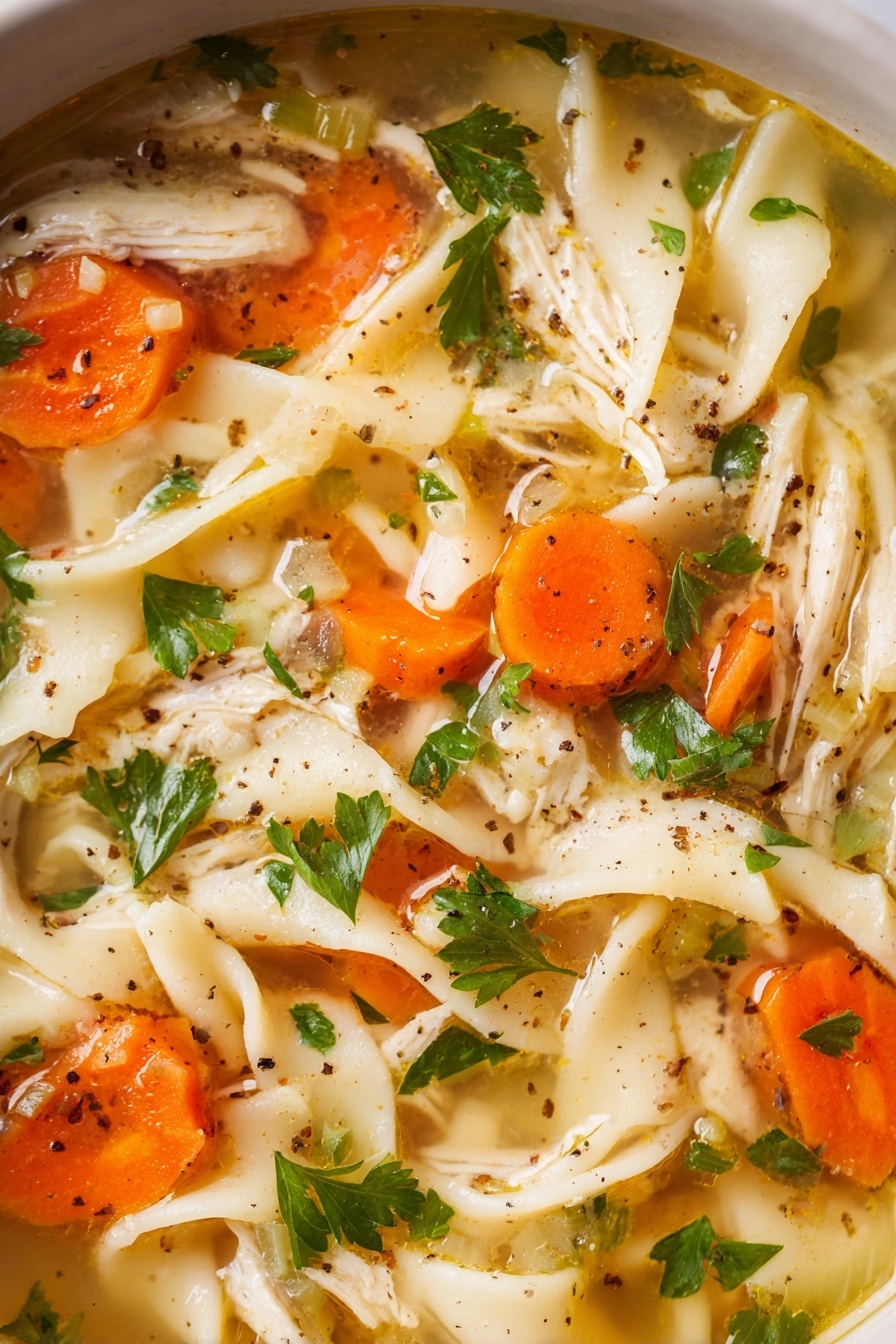 The image shows a close-up of chicken noodle soup with several layers visible. The first layer is a clear, light golden broth with a slight shine. Floating in the broth are wide, flat egg noodles that are creamy white and slightly soft in texture. Scattered throughout are large pieces of orange carrot slices and small chunks of white chicken. Bright green parsley leaves are spread on top, adding a fresh look. There are also small black pepper specks sprinkled across the soup. The entire scene is set on a white marbled surface. photo taken with an iphone --ar 2:3 --v 7