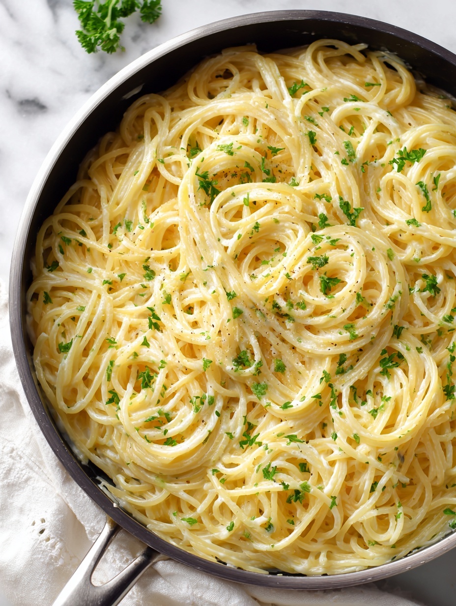The image shows a pan filled with creamy spaghetti pasta. The pasta is light yellow and coated evenly in a smooth sauce. Small bits of fresh green herbs are scattered throughout, adding small pops of color. Two silver forks are twirling some of the pasta from the pan, creating a motion effect. The pan sits on a white marbled surface with a soft white cloth partially visible beside it. The overall look is fresh and inviting, with the creamy sauce and herbs clearly visible on the spaghetti photo taken with an iphone --ar 2:3 --v 7