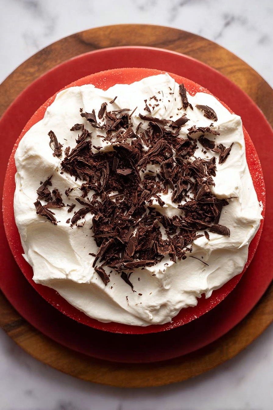 This dessert has three clear layers: the bottom layer is dark brown and crumbly, like a cookie crust, the middle layer is a thick, smooth, bright red jelly filling that looks shiny and firm, and the top layer is a fluffy white whipped cream spread evenly with some texture, sprinkled with small, thin dark chocolate shavings scattered on top. The dessert sits on a thick round wooden board with a white marbled background behind it. Photo taken with an iphone --ar 2:3 --v 7