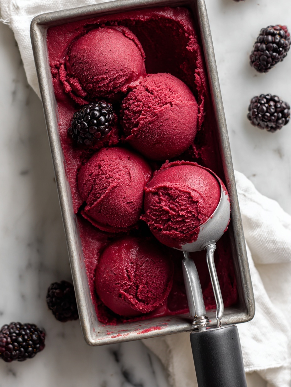A grey metal loaf pan filled with dark red blackberry sorbet, showing three rounded scoops carefully shaped inside it, with a silver ice cream scoop holding one scoop on the right side. Several plump blackberries are placed both inside the pan and scattered on a white marbled surface around the pan. The sorbet surface looks smooth and slightly shiny, with some texture showing where scoops were made. The scene is bright and clean, with a white cloth beneath the pan. Photo taken with an iphone --ar 2:3 --v 7