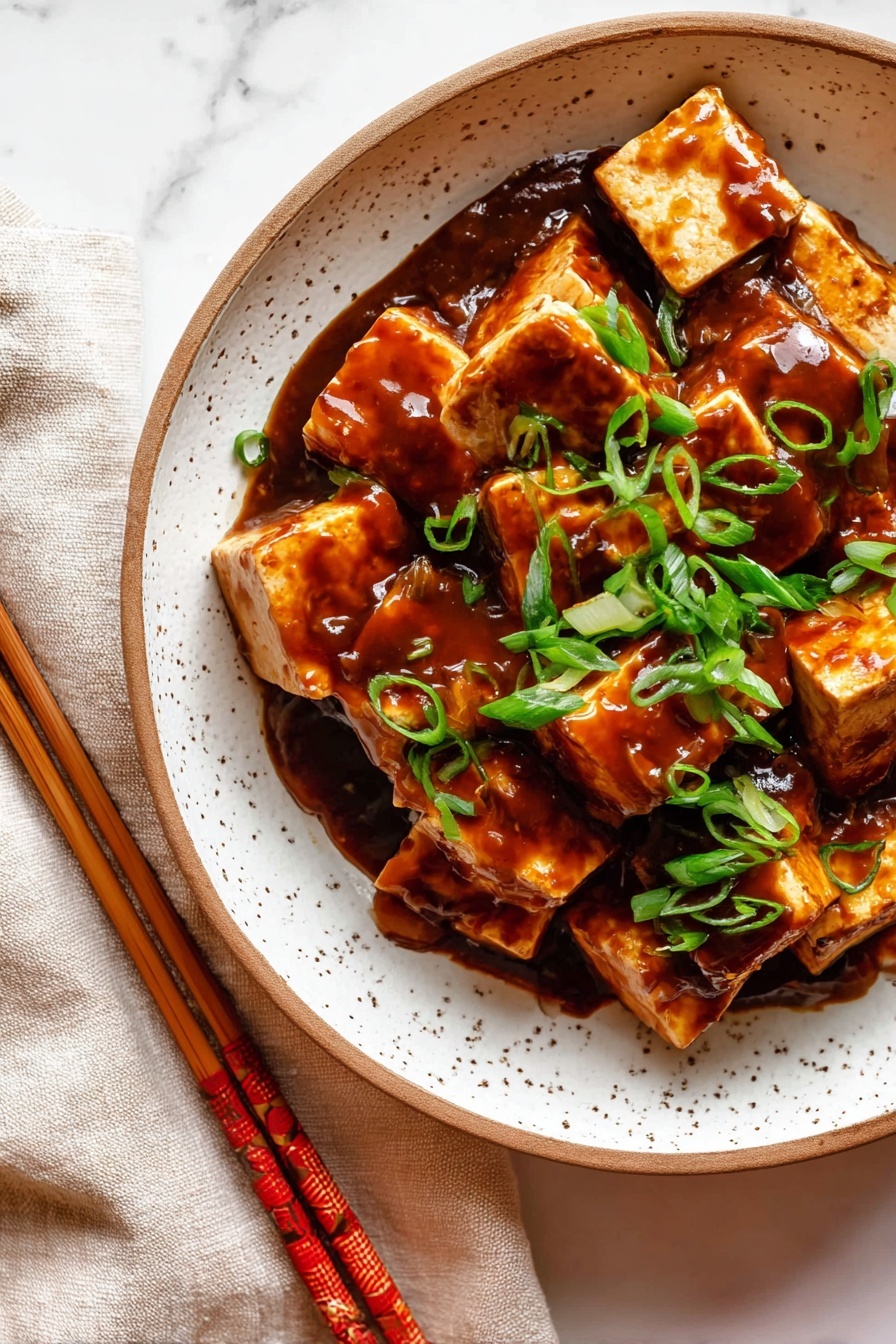 The dish shows a close-up of small, square pieces of brown tofu covered in a shiny, thick dark reddish-brown sauce. On top, there are bright green slices of fresh scallions scattered in an uneven layer. The food is served in a white speckled bowl with a brown rim, placed on a white marbled surface with part of a beige cloth and wooden chopsticks with red patterns visible to the side. Photo taken with an iphone --ar 2:3 --v 7