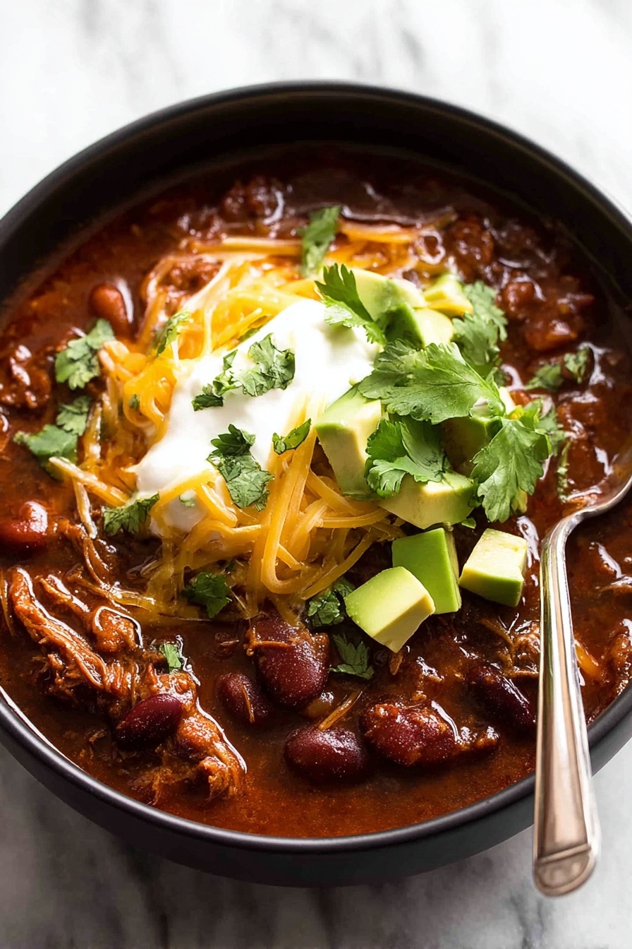 The image shows a black bowl filled with a thick, rich chili with visible red kidney beans and tender shredded meat in a dark red sauce. On top, there are orange shredded cheese, a dollop of smooth white sour cream, and chunky light green avocado pieces sprinkled with bright green chopped cilantro. A silver spoon rests inside the bowl on the right side, partially submerged in the chili. The background features soft focused green herb leaves on a white marbled surface. Photo taken with an iphone --ar 2:3 --v 7
