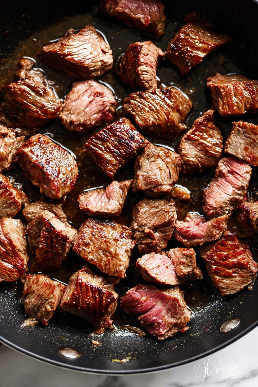 The image shows about twenty pieces of browned beef chunks cooking in a dark pan. Each piece is seared on the outside with a rich brown color, while some parts show a pinkish inside, indicating they are still cooking. The beef pieces vary in size and shape, irregular and thick, positioned spread across the bottom of the pan with a slight shine from cooking juices. The pan surface has little oil and bits of cooked residue, adding texture to the scene. The overall look is warm and hearty, with the meat appearing tender and juicy. Photo taken with an iphone --ar 2:3 --v 7