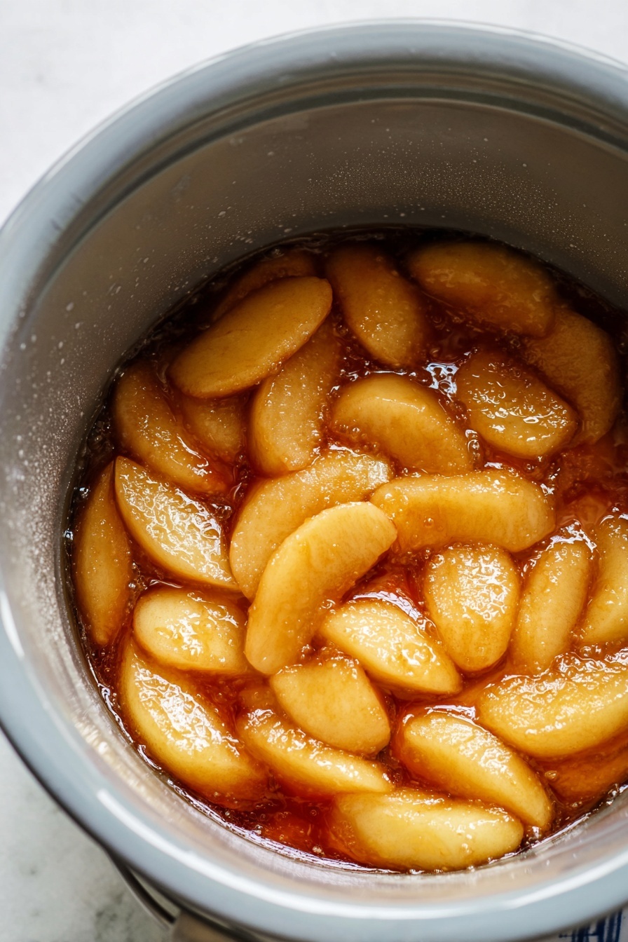 The image shows a close-up of a white round pot filled with a single layer of peeled, sliced apples covered in a shiny, thick caramel-colored syrup with visible cinnamon specks, giving the mixture a glossy, syrupy texture. The apples are soft and slightly translucent, arranged in a way that they touch each other but remain distinct pieces. The inside surface of the pot is smooth and light gray, contrasting with the warm tones of the apple mixture. The pot sits on a white marbled texture surface. Photo taken with an iphone --ar 2:3 --v 7